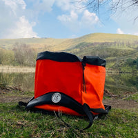 Orange Terrapin outdoor swimming bag on the ground with water in the background and hills in the distance. It is a sunny day.