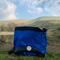 Blue Terrapin outdoor swimming bag on the ground with water in the background and hills in the distance. It is a sunny day.