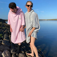 Two people near the water, one smiling at to the camera, both wearing Swim Feral organic towelling ponchos